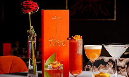 A selection of cocktails and drinks displayed alongside a red rose and orange gift box on a dark bar setting.