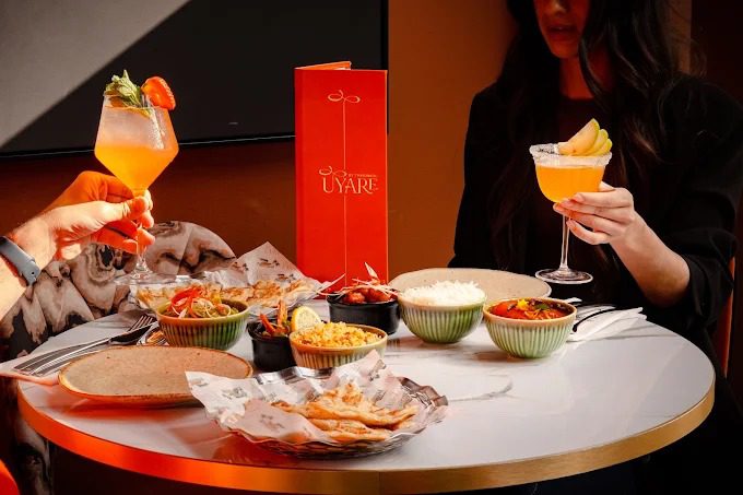 Two people toasting with cocktails at a table set with sharing plates and a red Uyaré menu, illustrating a romantic pre-theatre dinner for a special occasion.