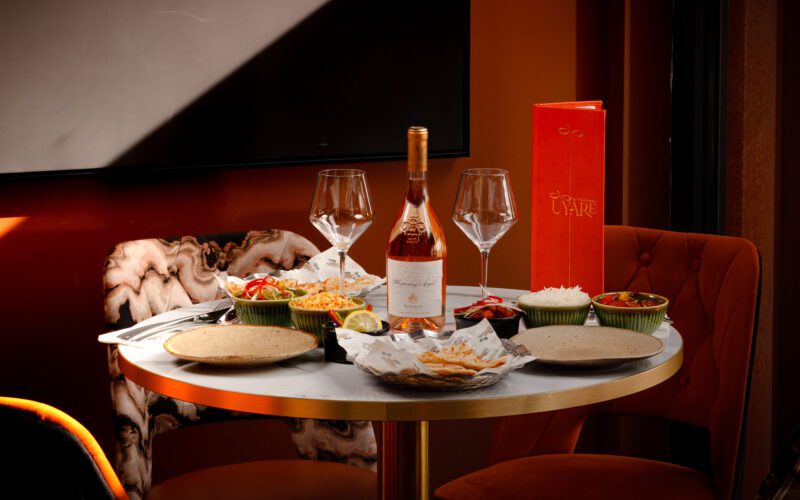 A round marble table set for two at Uyaré restaurant, with a bottle of rosé wine and two filled wine glasses, a red menu, and several sharing plates of tapas including flatbread, rice, and colourful dips, set against a dimly lit orange-toned dining area with plush velvet chairs.