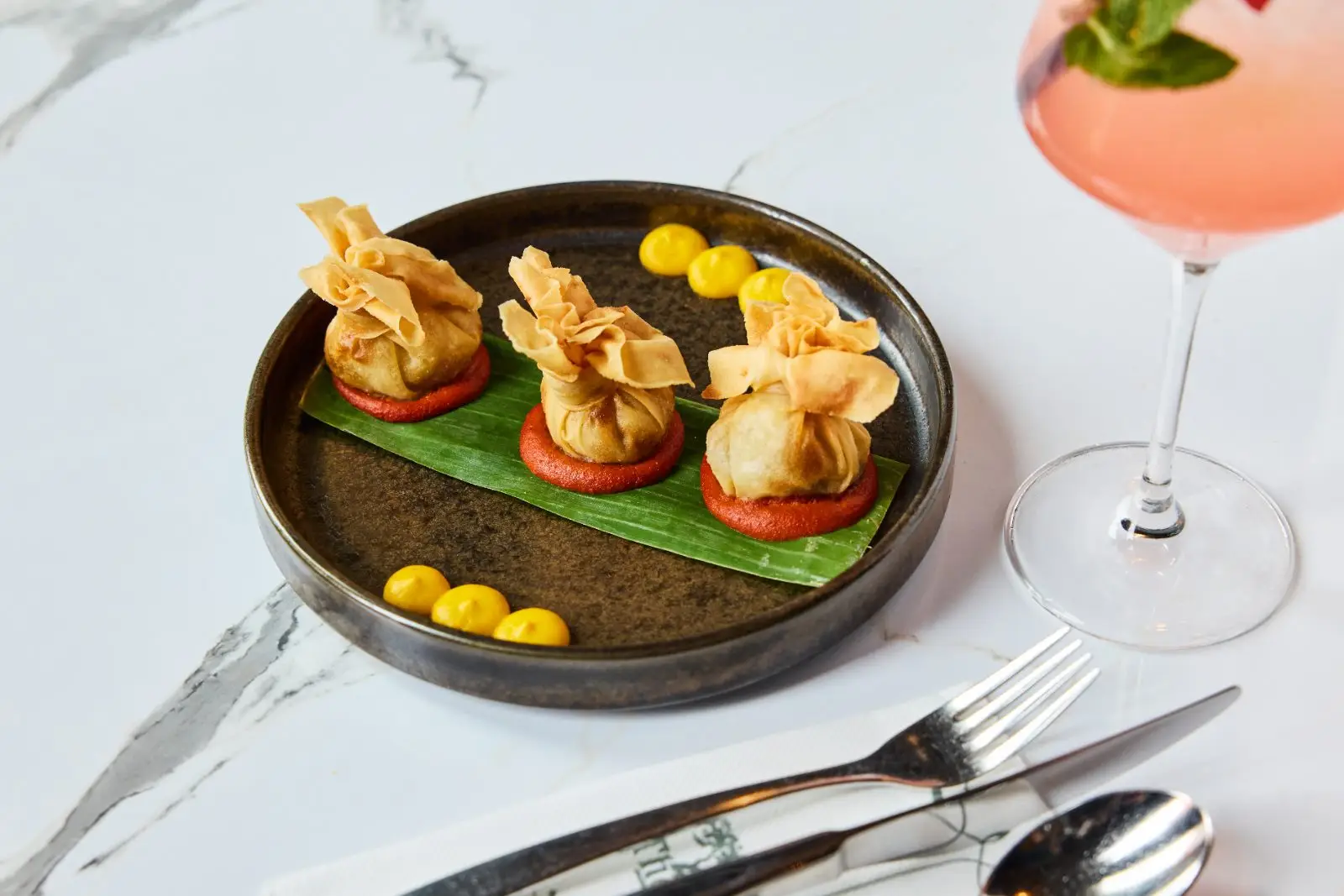 Three crispy pastry parcels on a banana leaf with a pink cocktail at UYARE