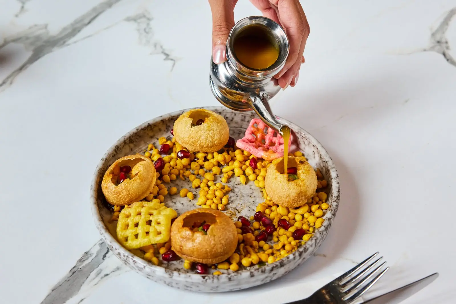 Spiced water being poured into crisp puri shells topped with pomegranate at UYARE